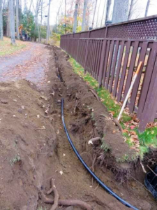 The ground is trenched along the side of the road next to a brown wooden fence. Conduit is in the ground.